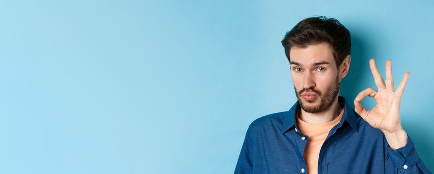 Image of silly young man pucker lips and show okay gesture, praise good thing, compliment excellent choice, standing on blue background.