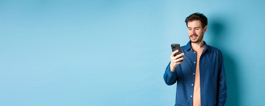 Handsome modern guy using mobile phone, reading smartphone screen and smiling, networking on blue background.