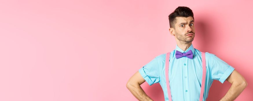 Image of young man with funny moustache and bow-tie standing in confident pose, staring with disbelief and doubts at camera, posing over pink background.