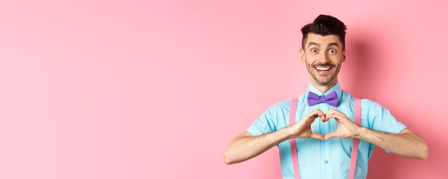 Happy Valentines day. Smiling funny guy with moustache showing heart sign and looking at lover, saying I love you, express feeling, standing over romantic pink background.