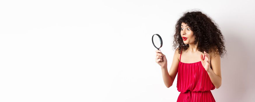 Excited woman in red dress look through magnifying glass and smiling, found interesting promo, standing on white background.