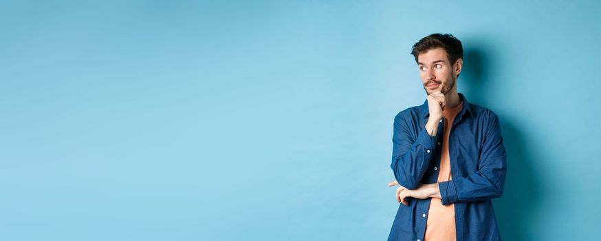 Thoughtful caucasian man looking aside at empty space and thinking, standing pensive on blue background. Copy space