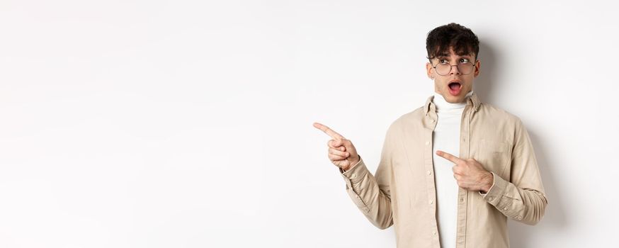 Amazed young man in glasses drop jaw, saying wow, pointing and looking left at awesome promo offer, stare with disbelief and impressed face, white background.