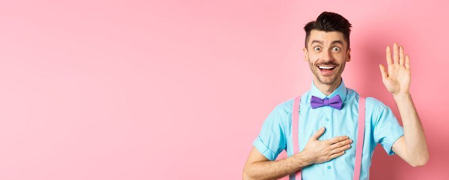 Happy young man telling truth, making promise, holding hand on heart and arm raised, swearing to be honest, smiling at camera, standing over pink background.