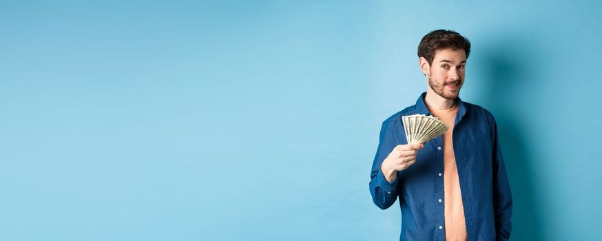 Unbothered rich guy showing cash and smiling, holding dollars in hand, standing on blue background. Copy space