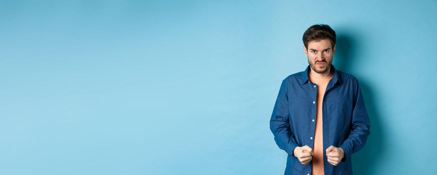 Angry and pissed-off man clench fists and stare with disdain at camera, trying to keep himself together, look with rage, standing on blue background.