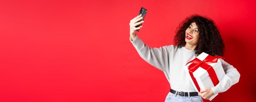 Holidays and tech concept. Happy woman taking selfie with her gift, holding present and smartphone, standing on red background.