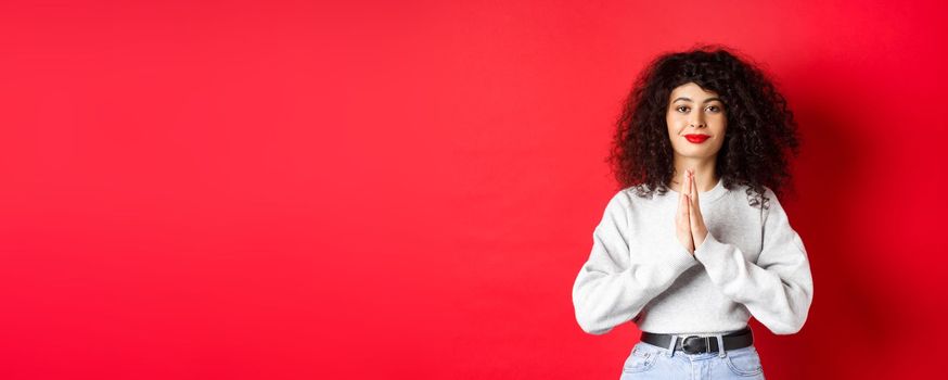 Namaste. Thankful young woman with curly hair and red lips show gratitude, smiling pleased at camera, standing on studio background.