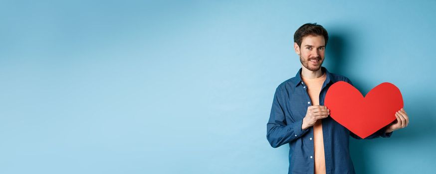 Happy man showing valentines heart and smiling, make romantic gift on lovers day, standing over blue background.