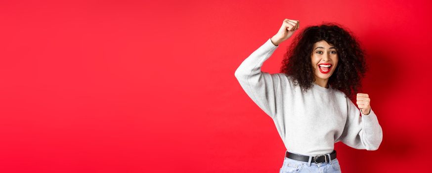 Cheerful young woman with curly hair, raising hand up and celebrating win, achieve goal or success, standing on red background.