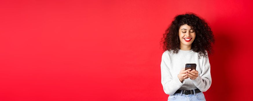 Happy positive girl chatting on phone, reading message and smiling, using social media app, standing on red background.