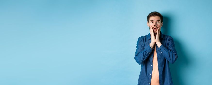 Portrait of guy gasping and dropping jaw with shocked face, staring at camera startled, standing on blue background.