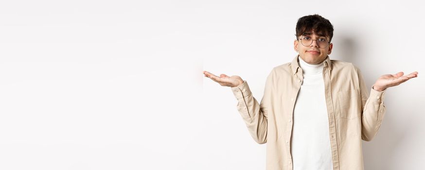 Portrait of handsome young man looking confused, know nothing, shrugging shoulders and spread hands sideways clueless, standing on white background.