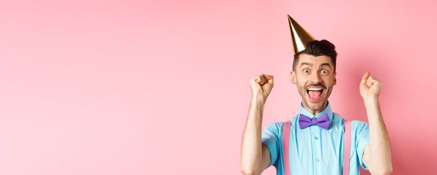 Holidays and celebration concept. Cheerful young man celebrating birthday party in cone hat and bow-tie, shouting yes with excitement and making fist pump gesture, pink background.