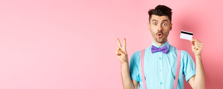 Shopping concept. Image of funny guy looking surprised while showing plastic credit card and victory sign, checking out promo deal, standing on pink background.
