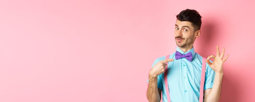 Image of young handsome guy in bow-tie and holiday outfit, pointing at himself and showing okay gesture, self-promoting on pink background.