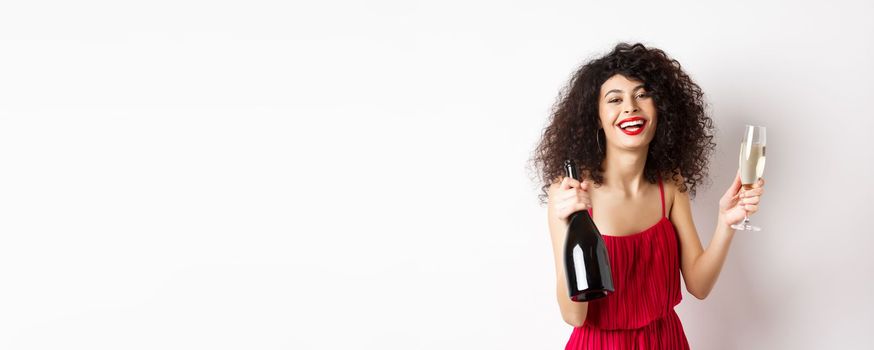 Happy party woman in red dress, laughing and holding bottle of champagne with glass, drinking and having fun, celebrating holiday, standing on white background.