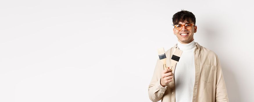 Hobbies and leisure concept. Stylish young man in glasses showing painting brushes and smiling, inviting to paint something, standing on white background.