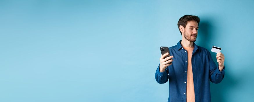 E-commerce concept. Man paying for online payment, reading credit card number and holding mobile phone, standing on blue background.