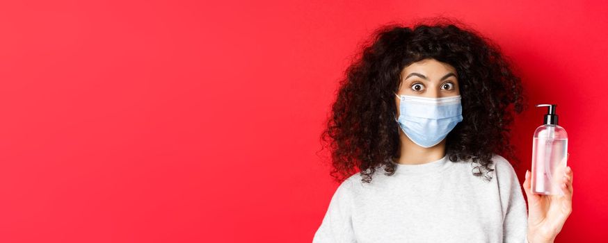 Covid-19, pandemic and quarantine concept. Excited girl with curly hair, wearing medical mask, showing bottle of hand sanitizer or antiseptic, standing on red background.