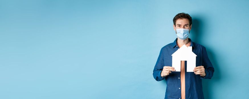 Real estate and quarantine concept. Happy young man in medical mask showing house cutout, buying property, standing on blue background.