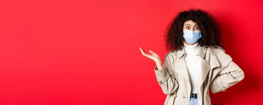 Covid-19, social distancing and quarantine concept. Shocked and confused european woman with curly hair, wearing medical mask from coronavirus, raise hand up puzzled, red background.