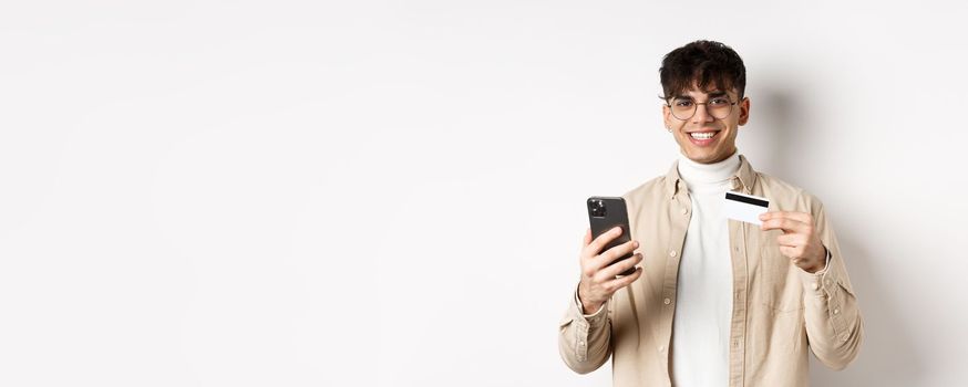 Portrait of natural young man in glasses paying in internet, showing smartphone and plastic credit card, standing on white background.