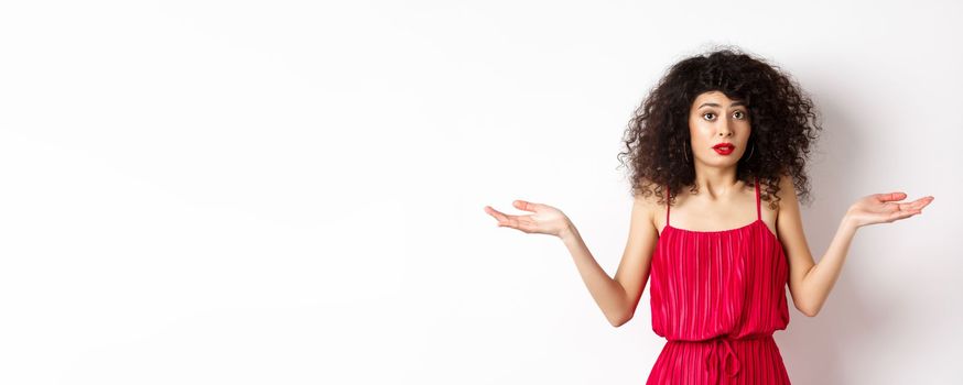 Confused lady in red dress shrugging shoulders, spread hands sideways and looking puzzled, know nothing, cant understand, standing over white background.