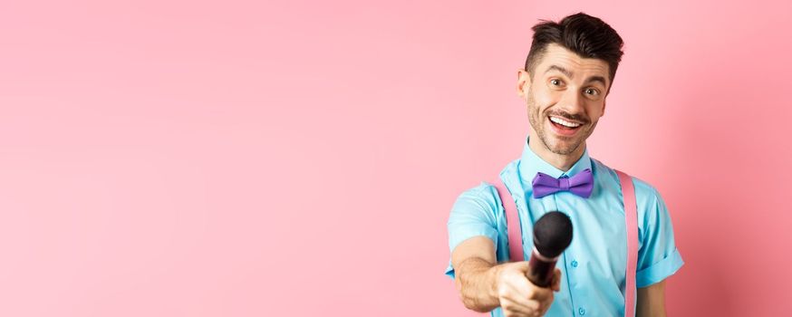 Smiling guy with moustache and bow-tie asking for comments or interview, stretch out hand with microphone, standing on pink background.