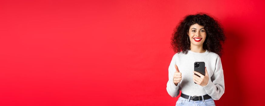 Smiling european woman looking happy and satisfied, using mobile phone and making thumb up, recommending application, standing on red background.