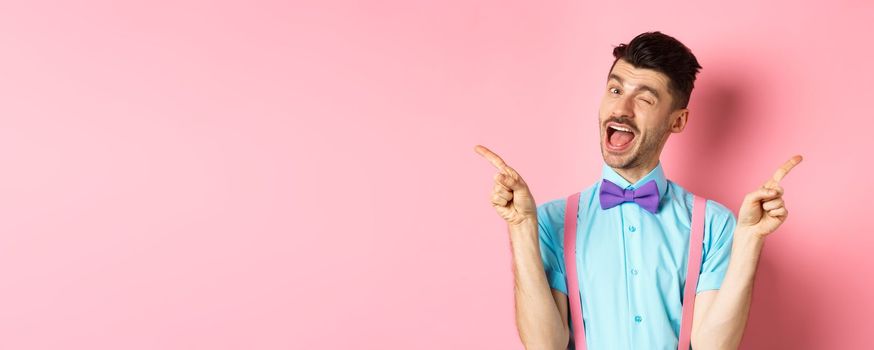 Cheerful young man smiling and winking, showing two sides, pointing fingers sideways and recommending promo offers on pink background.