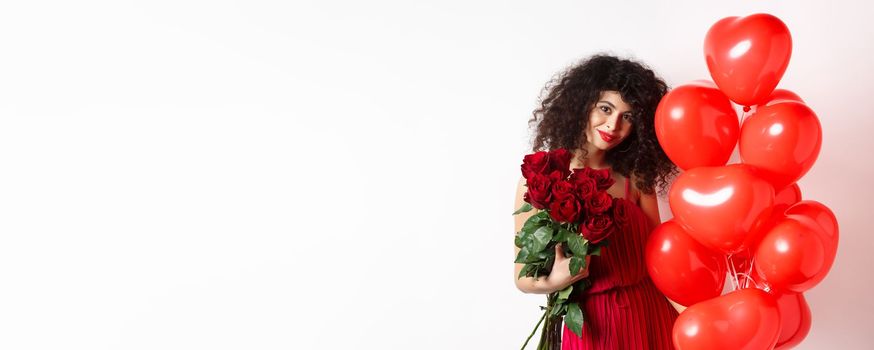 Gorgeous curly haired girlfriend in evening dress, having a date, holding red roses from boyfriend and posing near romantic hearts balloons, white background.