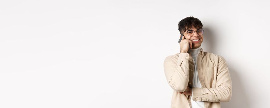 Real people concept. Happy young guy talking on mobile phone, look aside and holding smartphone near ear, standing on white background.