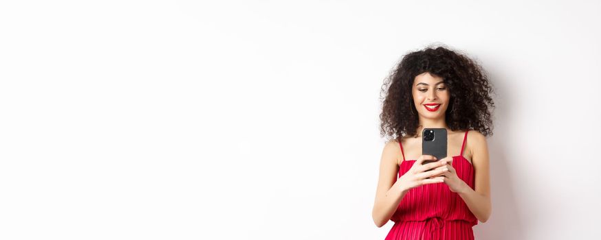 Happy fashionable woman taking photo on smartphone, photographing something on mobile and smiling, standing in red dress on white background.