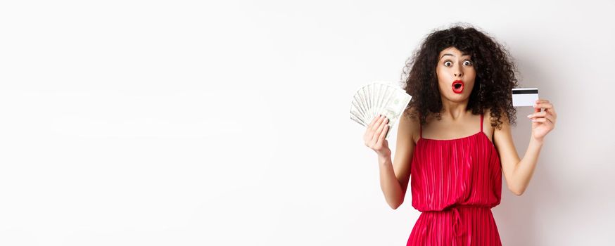 Impressed lady with curly hair and trendy red dress, saying wow, showing plastic credit card with cash, standing on white background.