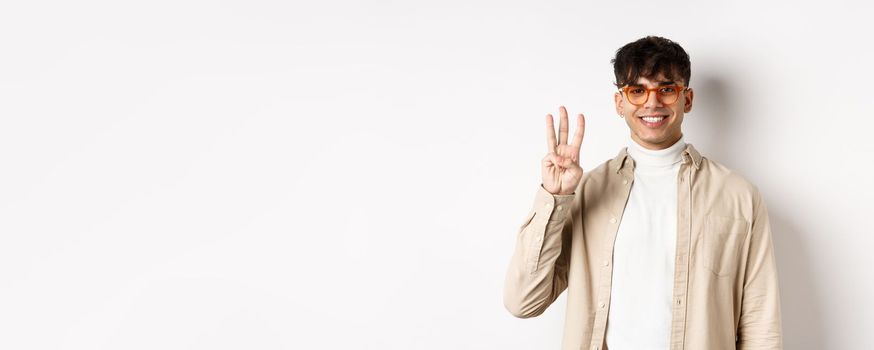 Handsome caucasian guy in glasses showing three fingers and smiling, standing on white background.