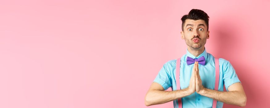 Young guy with cute face pucker lips and asking please, begging for favour, need something and lookin gpleading at camera, standing over pink background.