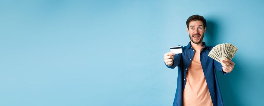 Cheerful caucasian guy extending hands with dollars and plastic credit card, smiling happy at camera, standing on blue background.