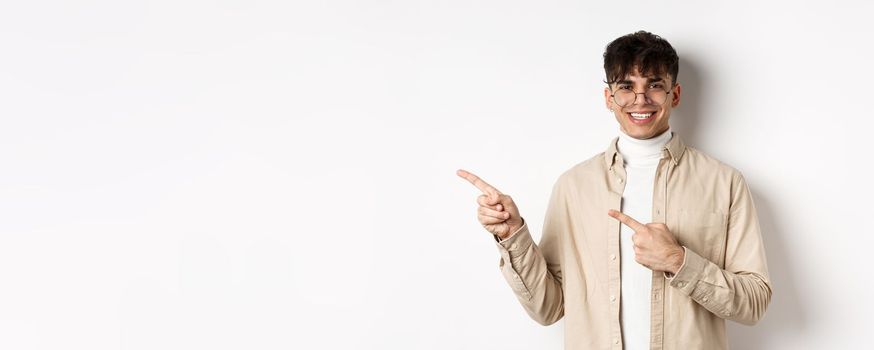 Portrait of handsome caucasian young man in glasses showing logo, smiling and pointing fingers left, inviting to check out promo deal, white background.