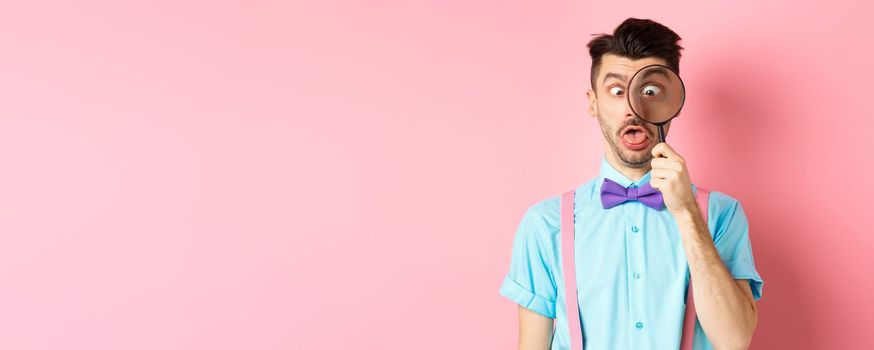 Funny man in bow-tie look through magnifying glass, squinting and making silly faces, standing on pink background.