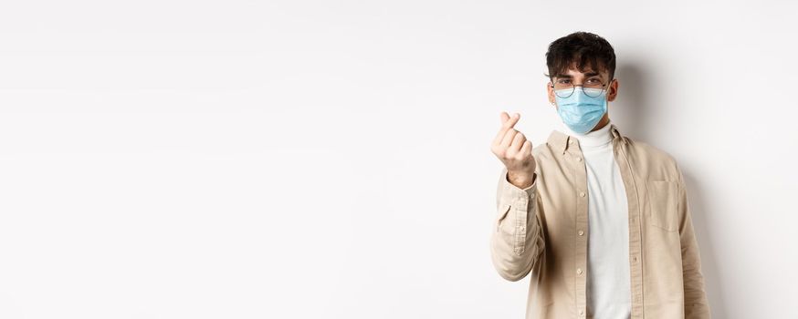 Covid-19, health and real people concept. Cute young man in medical mask showing finger heart and look at camera, standing on white background.