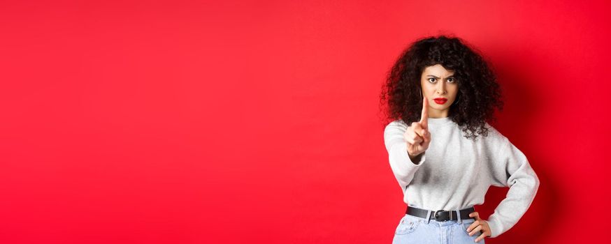 Serious confident woman say no, extend one finger to stop you, prohibit something bad, standing determined against red background.