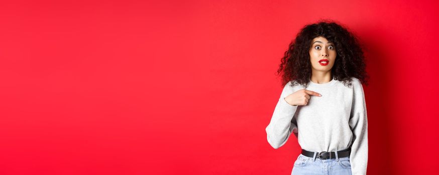 Surprised attractive female model pointing at herself, raising eyebrows and looking with disbelief at camera, standing on red background.