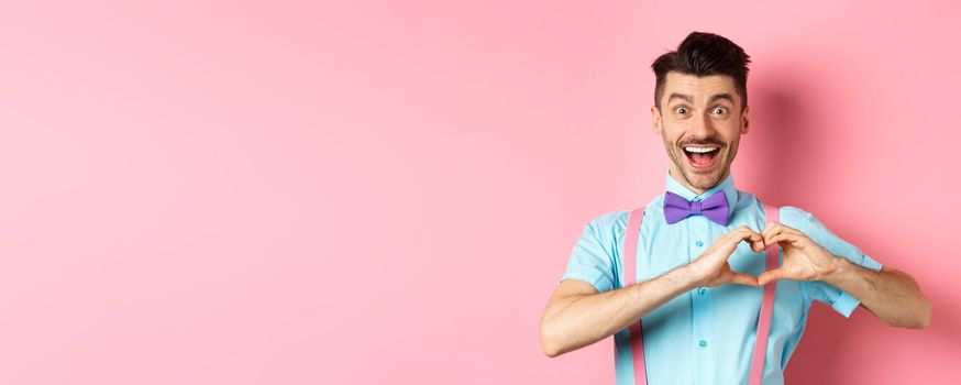 Excited smiling man showing pounding heart and looking with love, standing over romantic pink background. Valentines day concept.