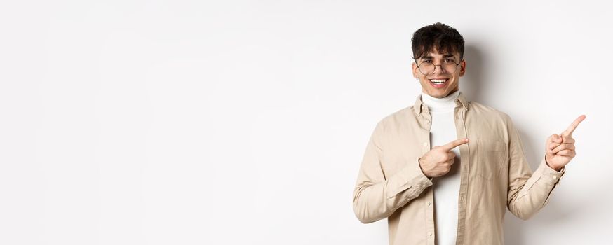 Proud and happy young man in glasses showing logo, pointing fingers at upper right corner and smiling, standing on white background.