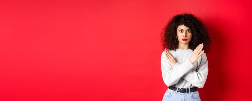 Serious caucasian woman with curly hair say no, making cross gesture to stop or prohibit something bad, standing displeased on red background.