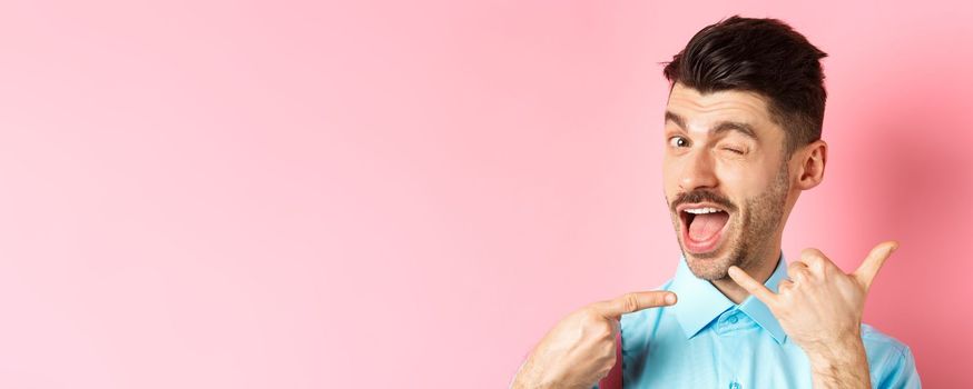 Cheeky and funny guy showing phone gesture, asking to call him and winking, telling dial his number, standing over pink background.