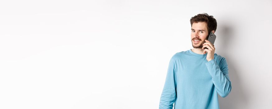 Technology concept. Young male model talking on mobile phone, calling someone on smartphone and smiling, standing over white background.