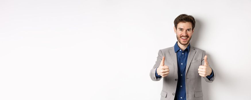 Cheerful and positive young man in business suit showing thumbs up and smiling, like something good, praise good job, standing happy on white background.