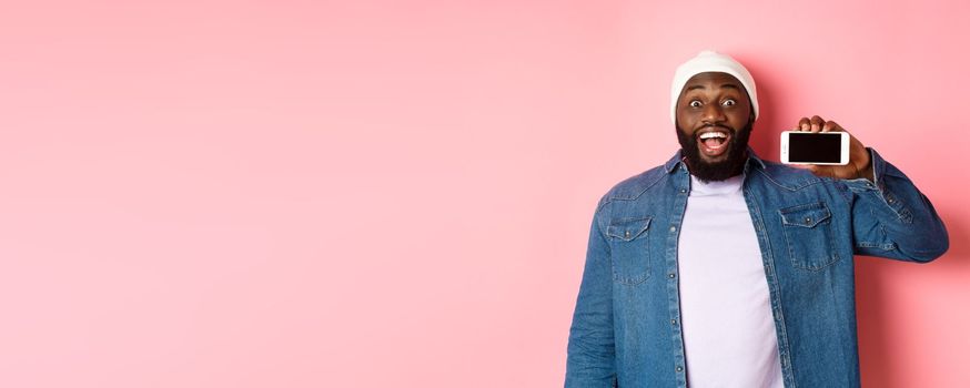 Online shopping and technology concept. Amazed Black man showing phone screen horizontally and staring at camera excited, standing over pink background.